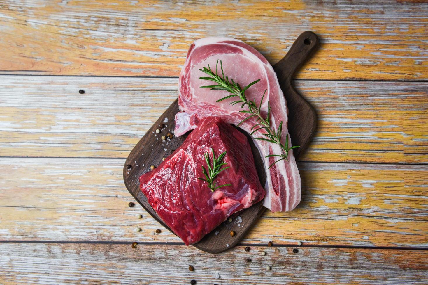 Carne fresca de ternera en rodajas y chuletas de cerdo hierbas especias romero sobre tabla de cortar de madera de fondo - bistec de ternera cruda foto