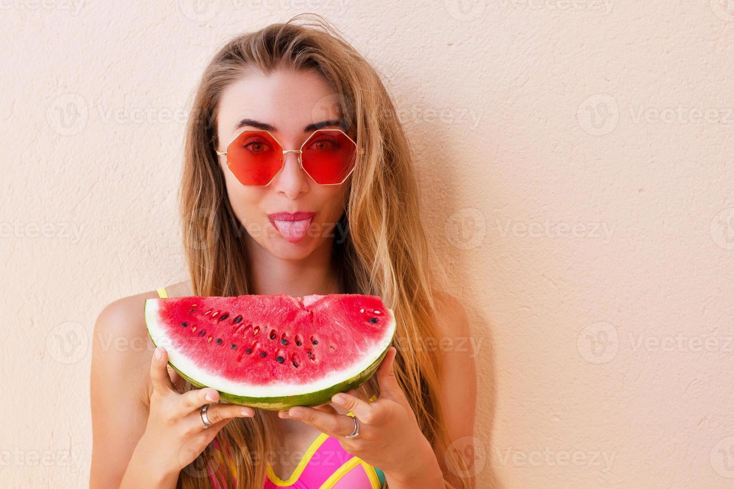 Primer plano de la moda bohemia joven y bella mujer rubia caucásica con gafas de sol rojas sosteniendo una rodaja de sandía. atractiva chica en retrato de pintura corporal traje de baño al aire libre. foto