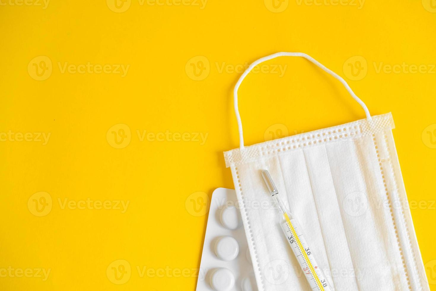 Mascarilla médica desechable, pastillas y un termómetro de mercurio sobre un fondo amarillo. foto