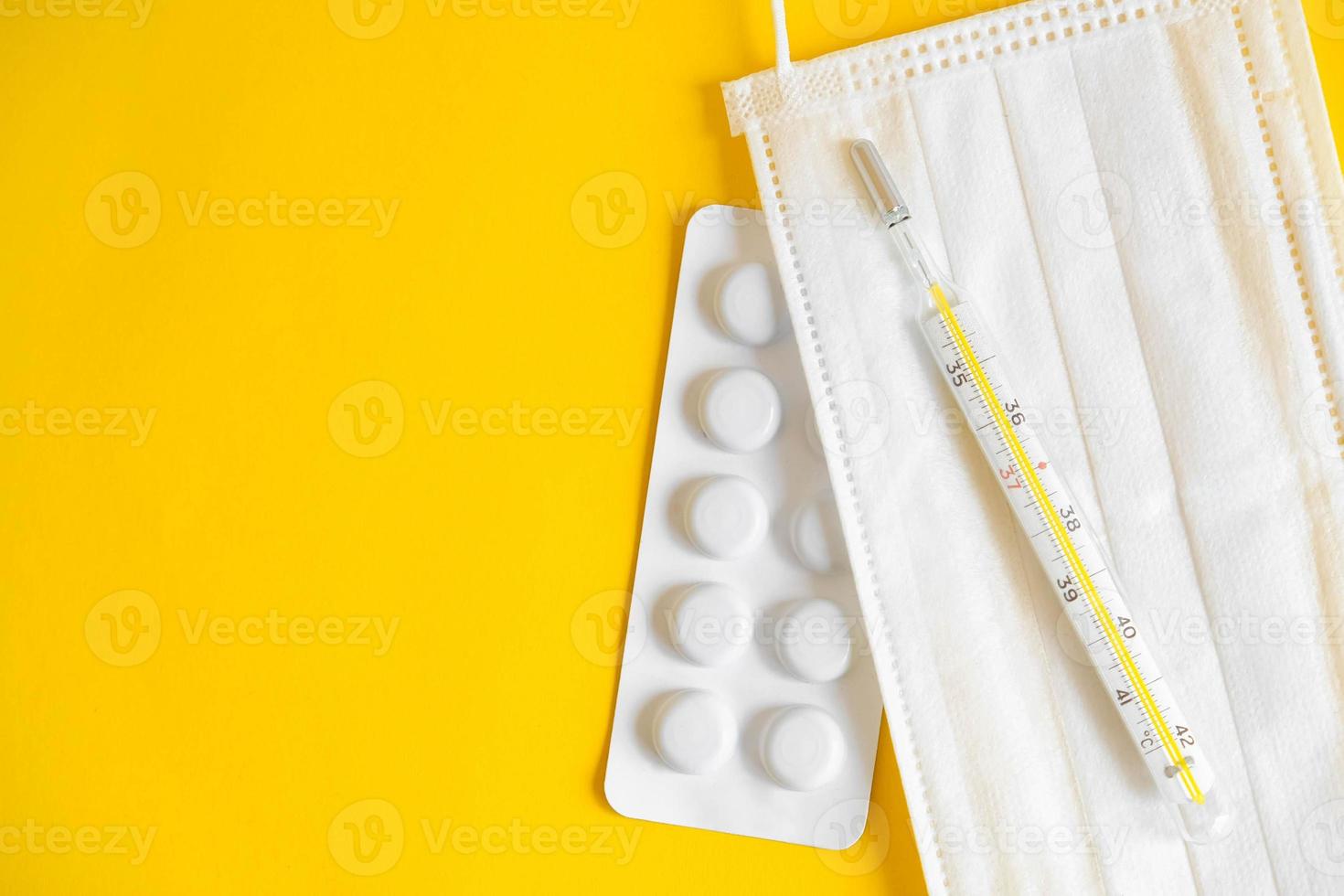 Mascarilla médica desechable, pastillas y un termómetro de mercurio sobre un fondo amarillo. foto