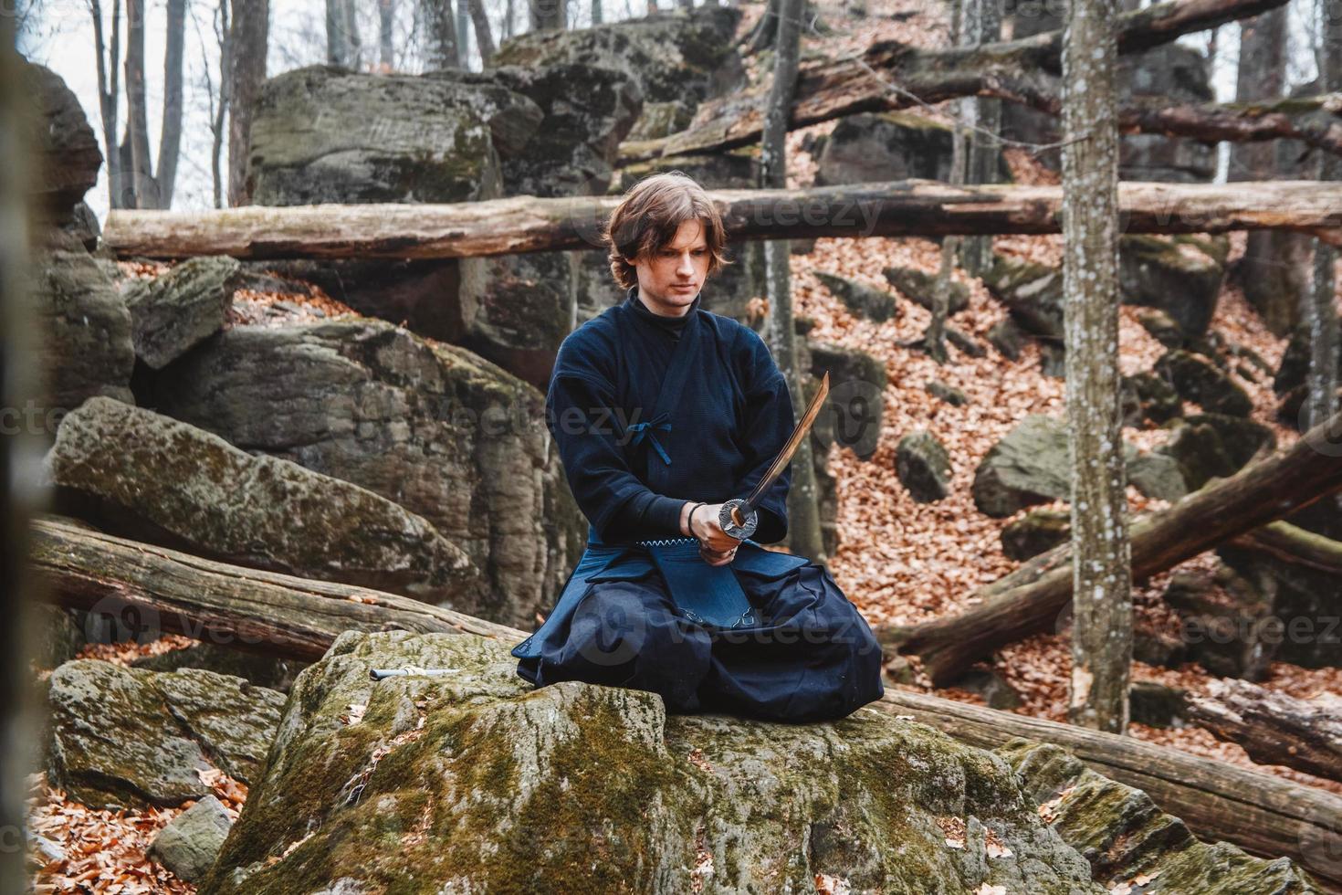 Man in black kimono with a sword meditates and concentrates on rocks and forest background photo