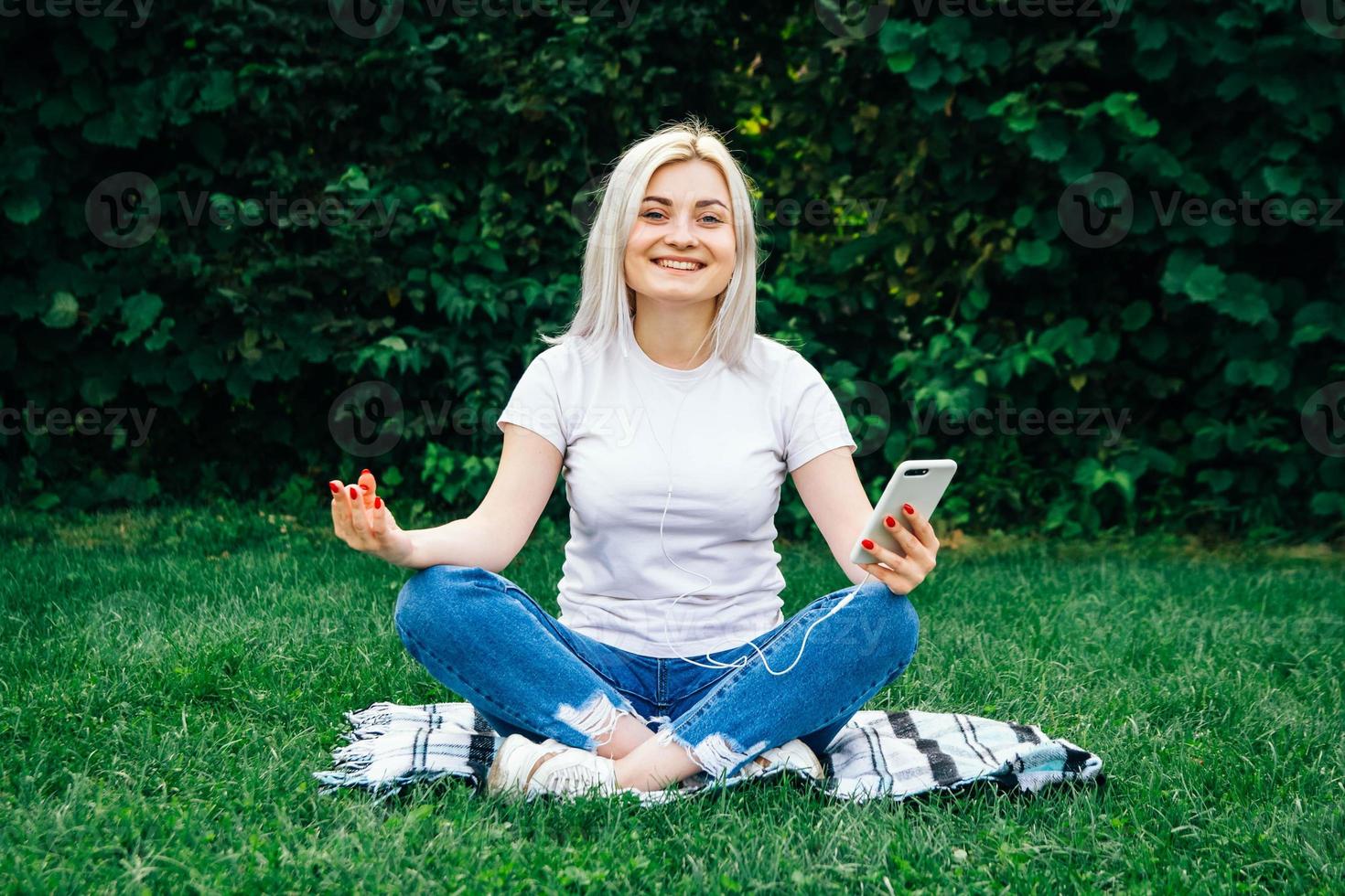 Woman in headphones and smartphone in hands sits in meditative pose listening to music on green grass photo
