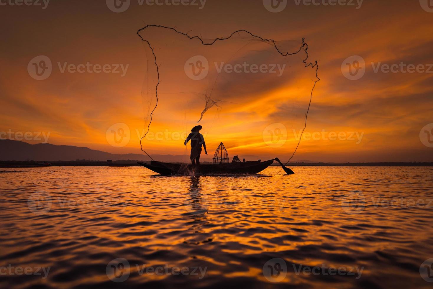 Pescador asiático en barco de madera echando una red para pescar peces de agua dulce en el río natural temprano en la mañana antes del amanecer foto