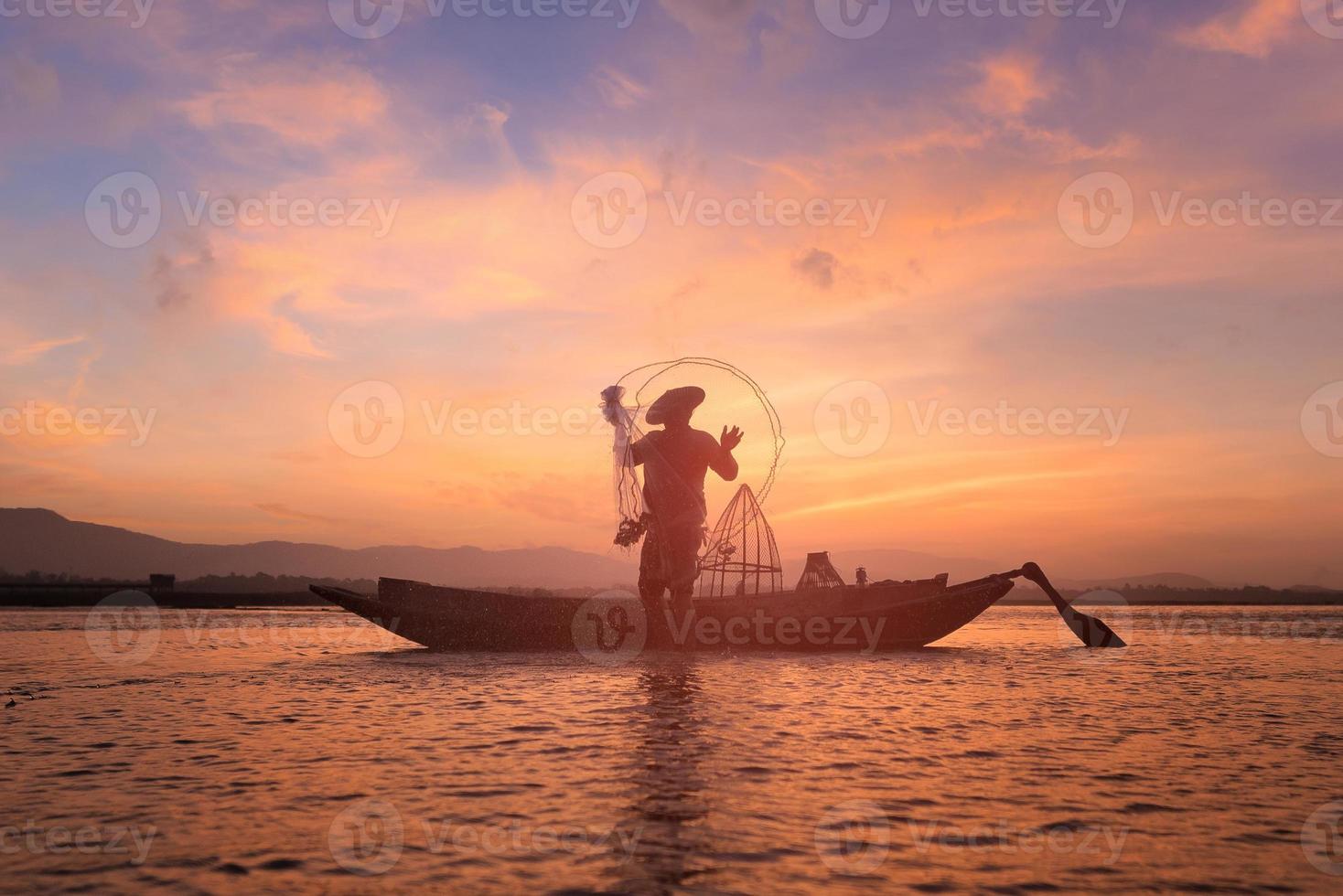 Asian fisherman on wooden boat casting a net for catching freshwater fish in nature river in the early morning before sunrise photo