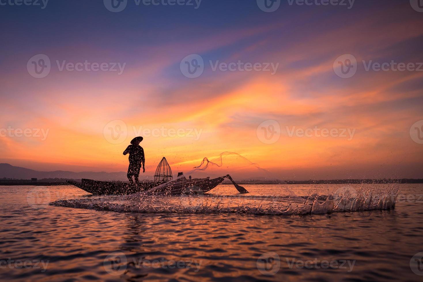 Asian fisherman on wooden boat throwing a net for catching freshwater fish in nature river in the early morning before sunrise photo