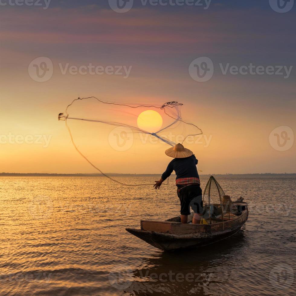 Asian fisherman on wooden boat throwing a net for catching freshwater fish in nature river in the early during sunrise time photo