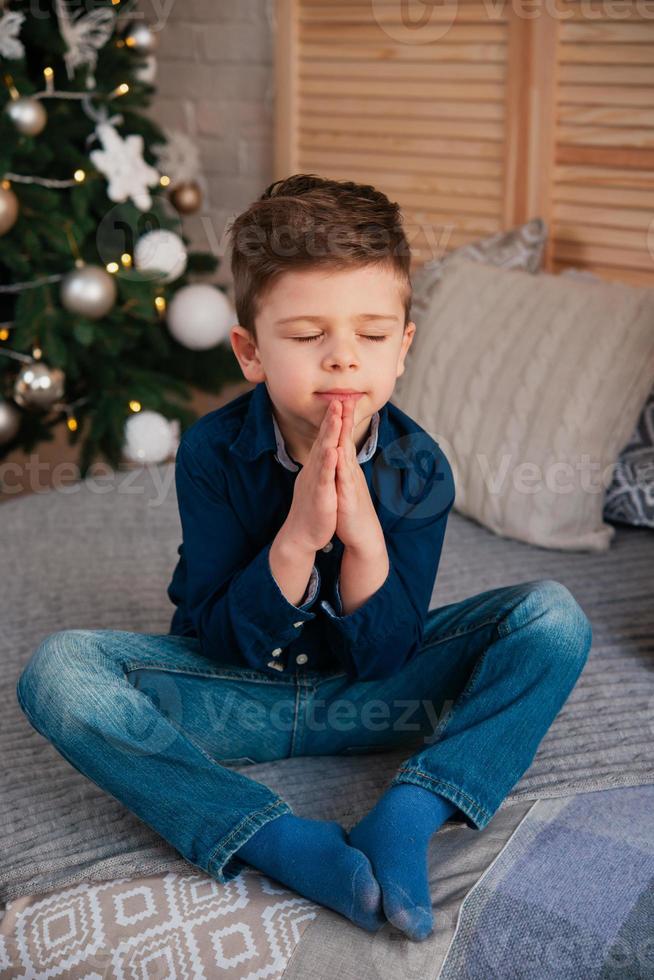 un pequeño niño dulce se sienta con los ojos cerrados cerca del árbol de año nuevo y pide un deseo, cruzando las manos cerca de su pecho foto