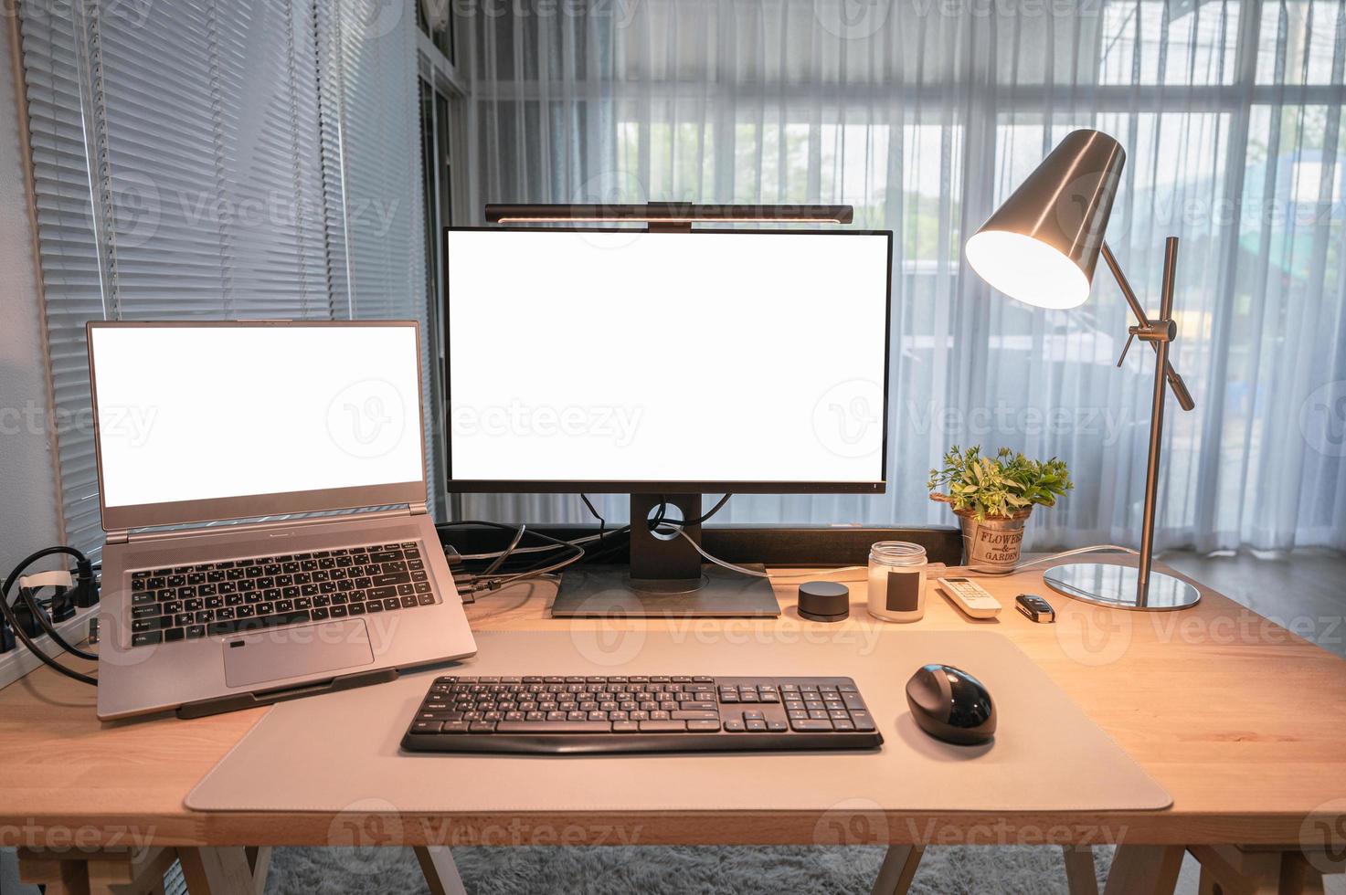 Computer device with blank monitor, laptop, table lamp, keyboard, mouse on wooden desk in home office photo
