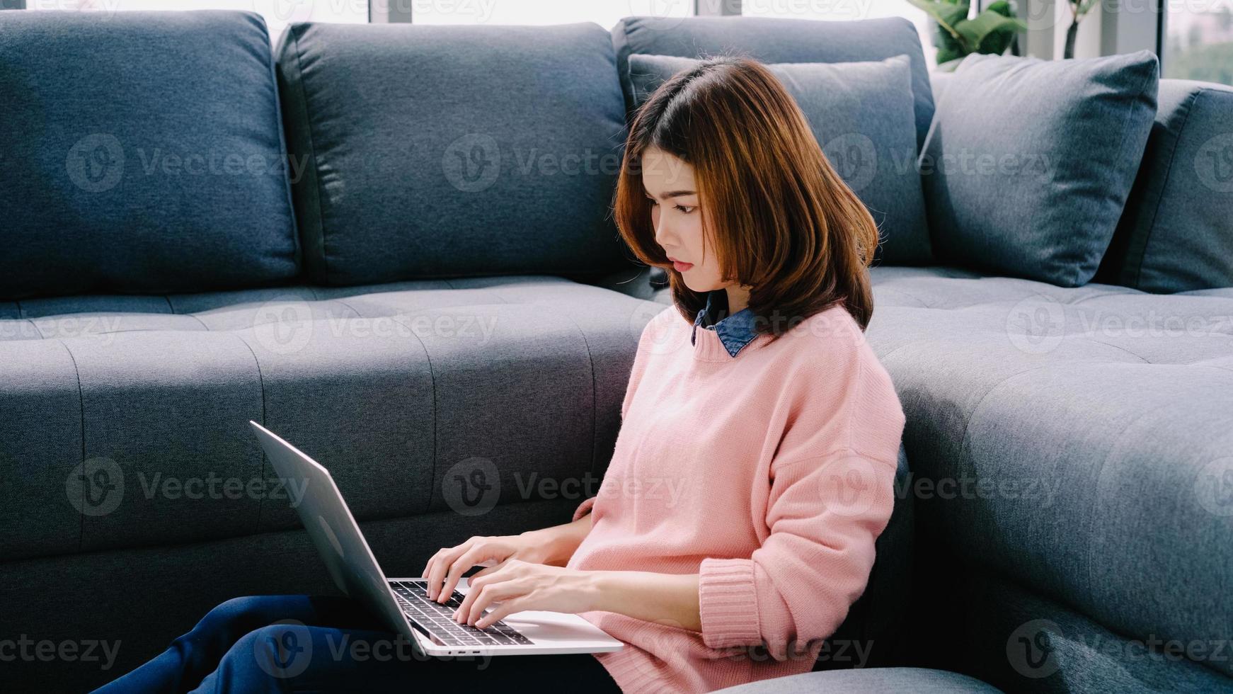 Young asian woman doing research work for her business. Smiling woman sitting on sofa relaxing while browsing online shopping website. Happy girl browsing through internet during free time at home. photo