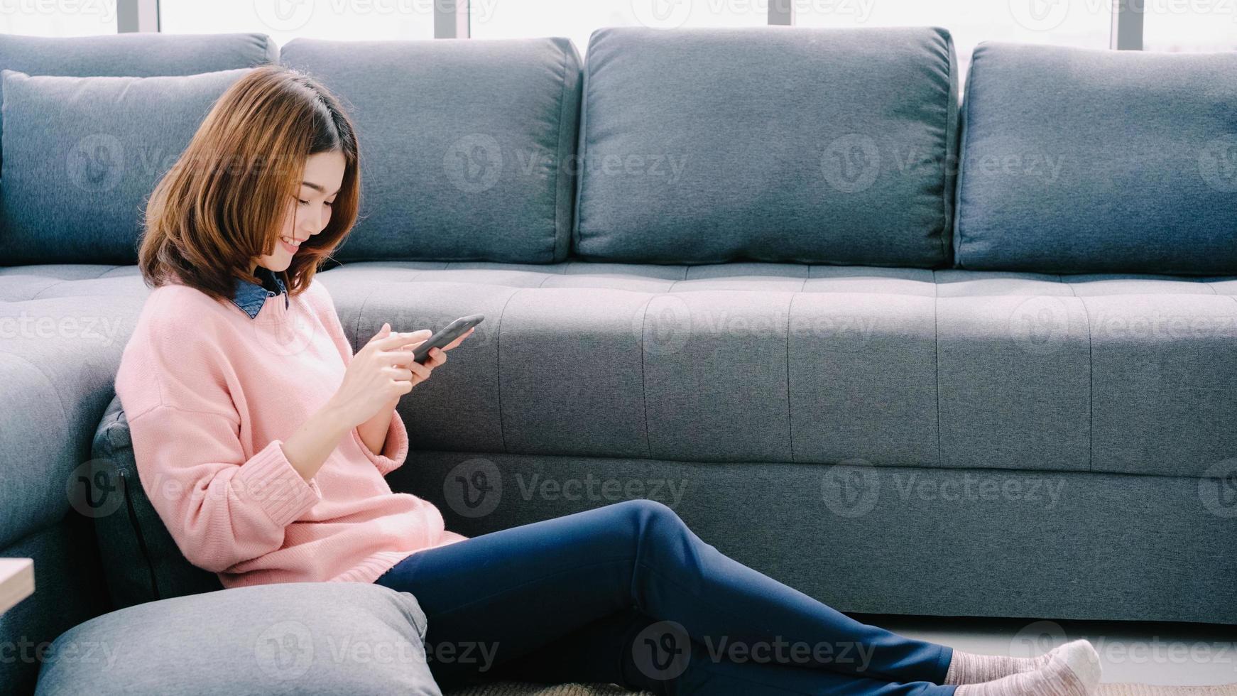 Asian woman using smartphone while lying on home sofa in her living room. Happy female use phone for texting, reading, messaging and buying online at home. Lifestyle woman at home concept. photo