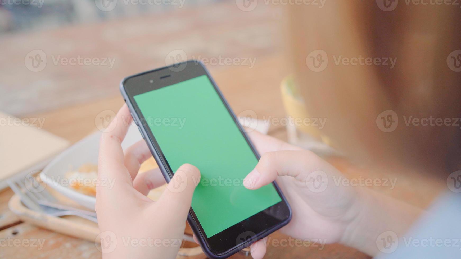 Young asian woman using black mobile phone device with green screen. Asian woman holding smartphone, scrolling pages while sitting in cafe. Chroma key. photo
