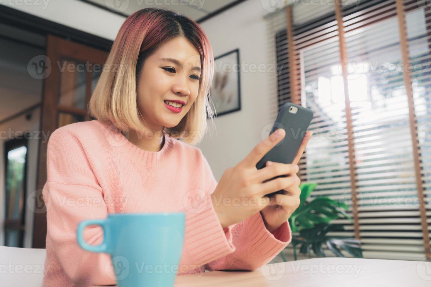 Young Asian woman using smartphone while lying on the desk in her living room. Happy female use phone for texting, reading, messaging and buying online at home. Lifestyle woman at home concept. photo