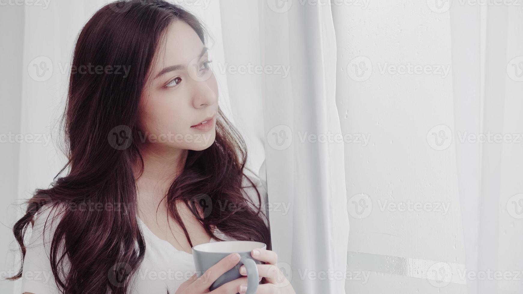 Young Asian woman enjoying her coffee or tea near the wet window in her bedroom at home. Beautiful teenager female drinking beverage and relax in rainy day. photo