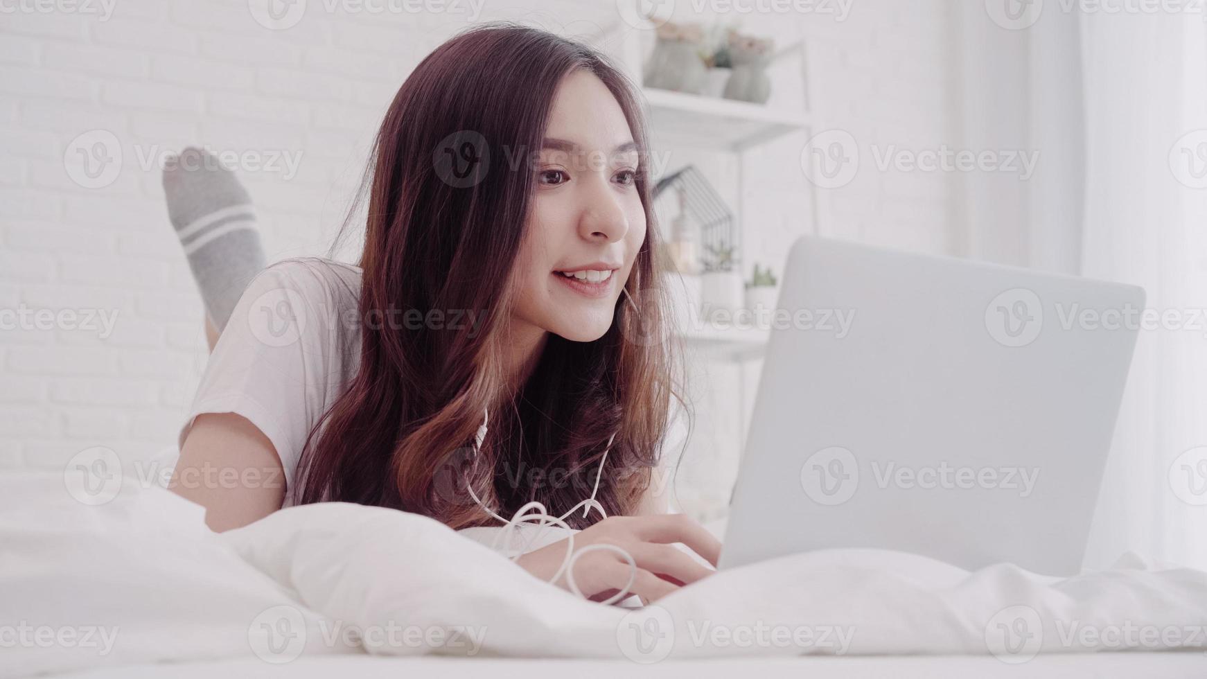 Portrait of beautiful attractive Asian woman using computer or laptop and listening music while lying on the bed when relax in her bedroom at home. Lifestyle women using relax time at home concept. photo