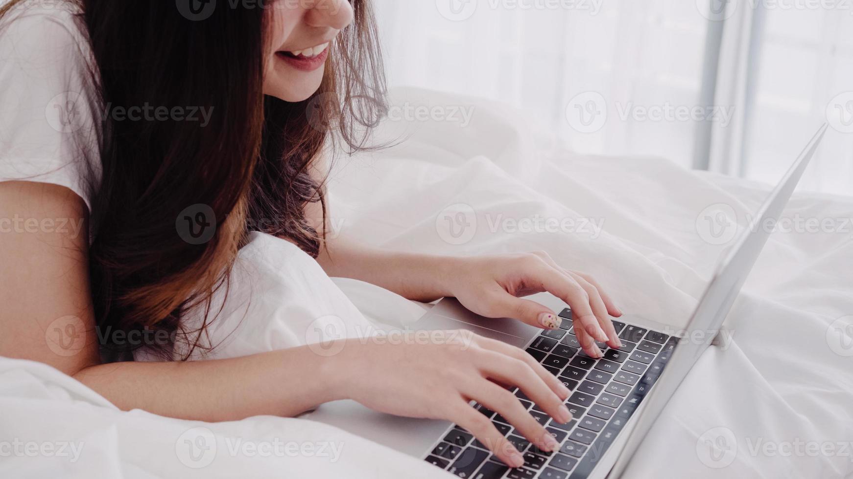 Portrait of beautiful attractive Asian woman using computer or laptop and listening music while lying on the bed when relax in her bedroom at home. Lifestyle women using relax time at home concept. photo