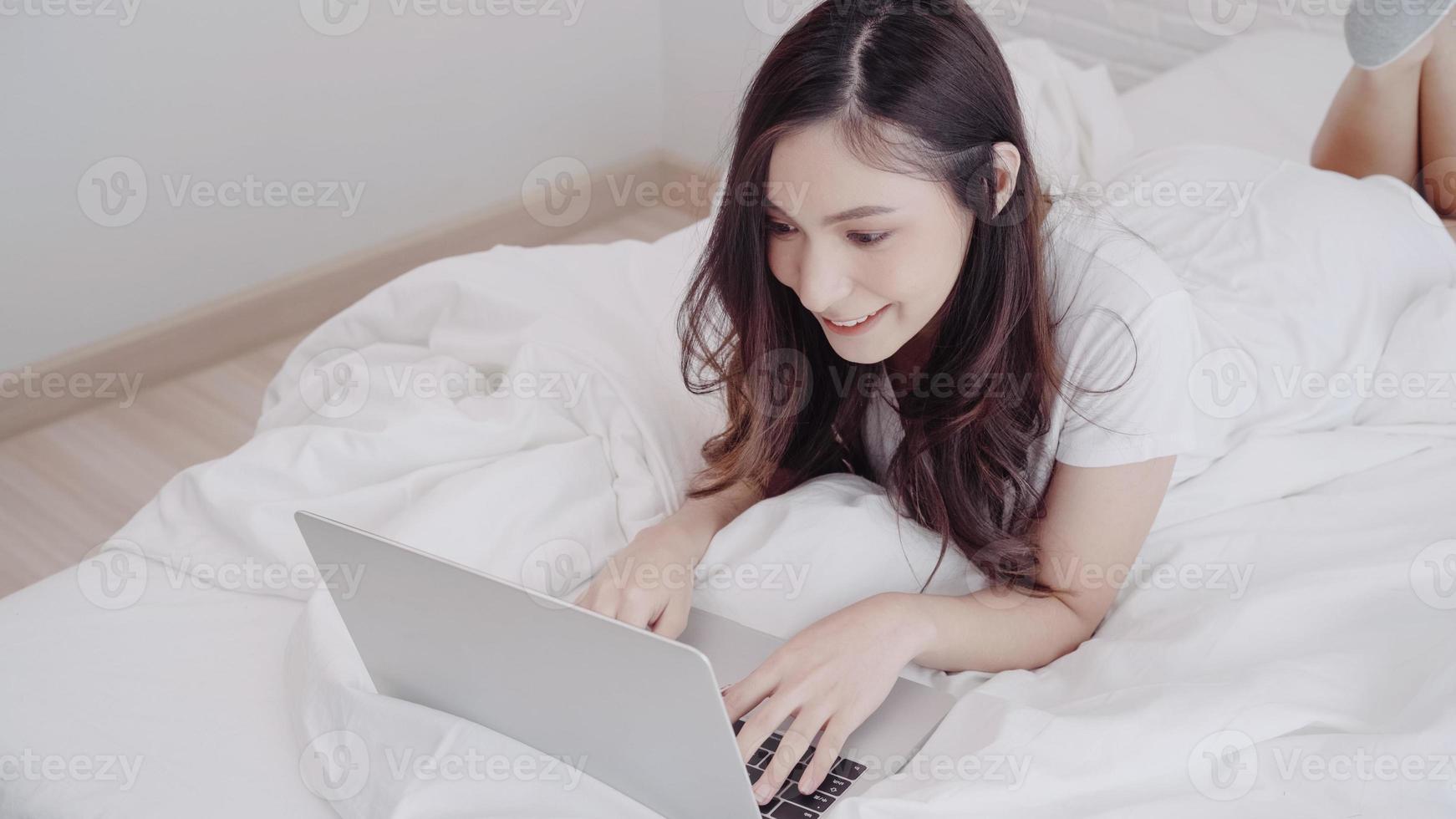 Portrait of beautiful attractive Asian woman using computer or laptop and listening music while lying on the bed when relax in her bedroom at home. Lifestyle women using relax time at home concept. photo