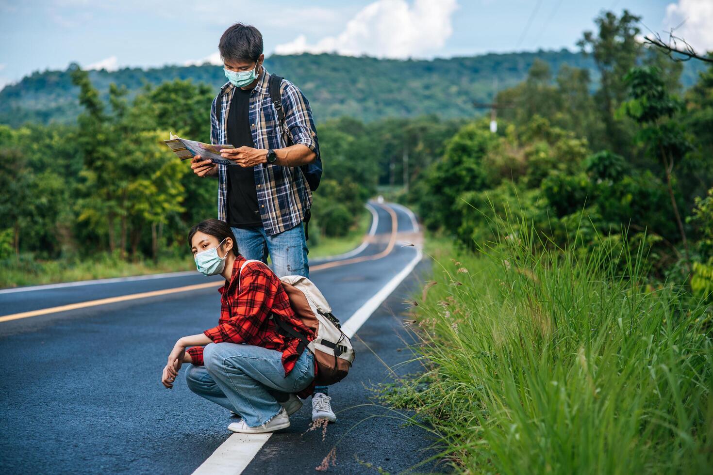 Female tourists sitting, male tourists looking at the map, both wearing masks and on the side of the road. photo