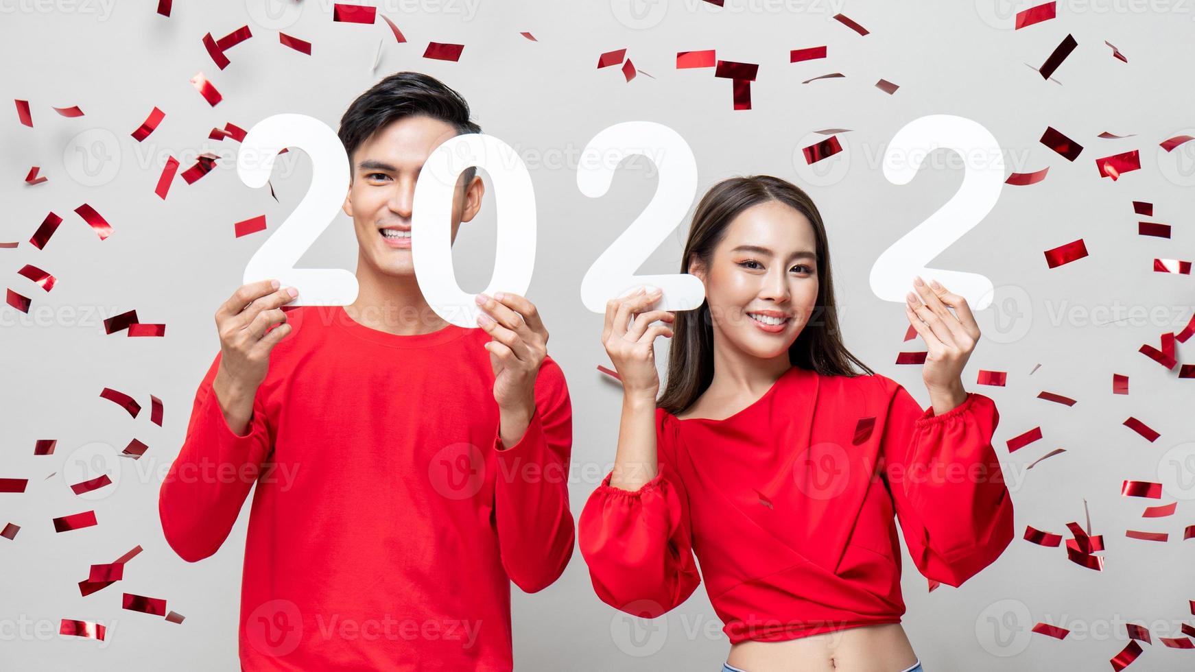 Lovely Asian couple smiling and showing number 2022 for new year concept on light gray studio background with confetti photo