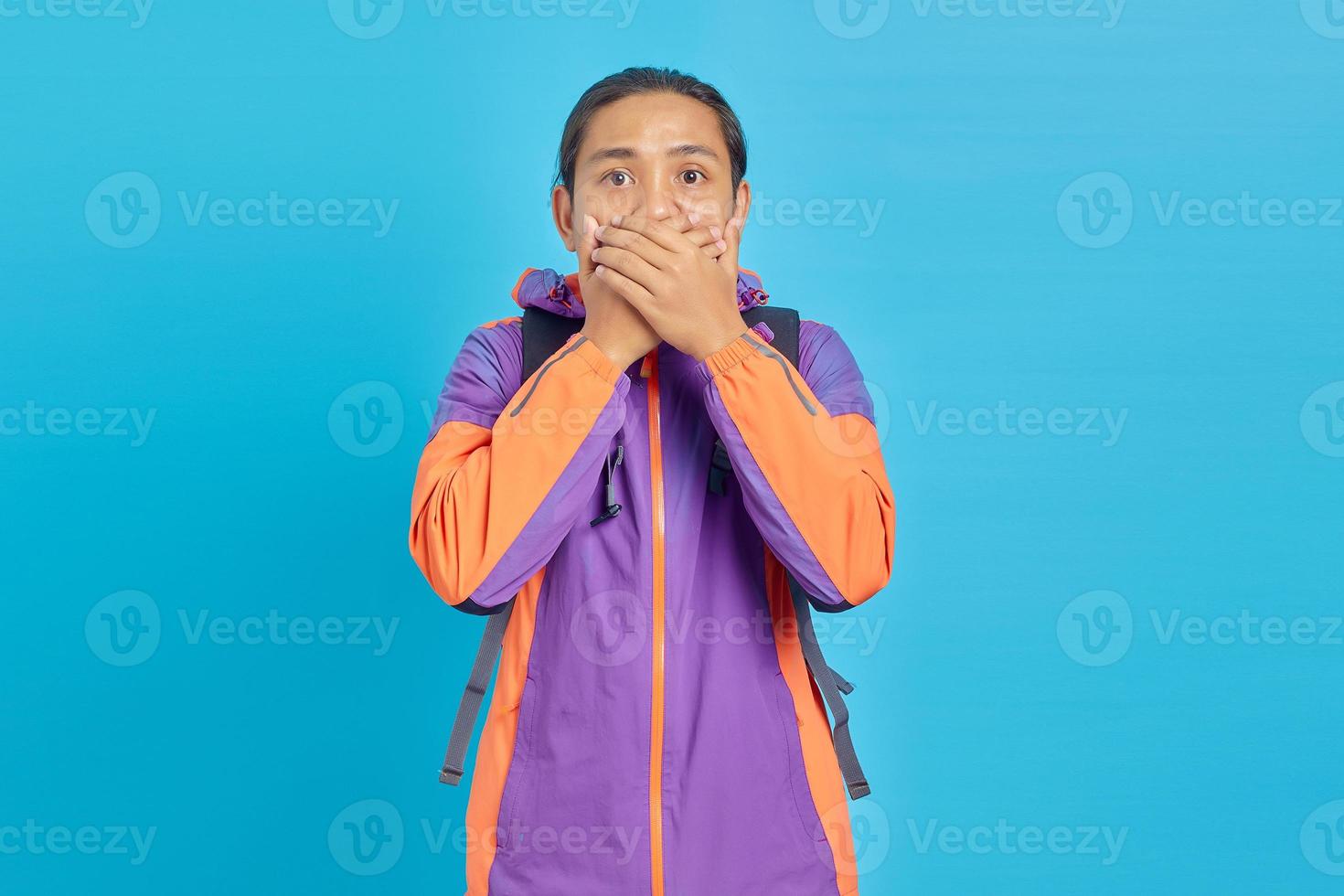 Conmocionado joven asiático cubriendo la boca con la mano sobre fondo azul. foto
