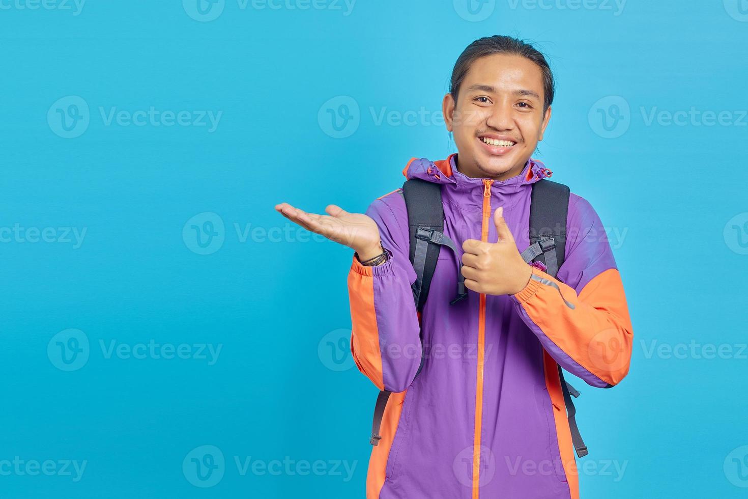Portrait of smiling young Asian man showing palm hands and thumbs up on blue background photo