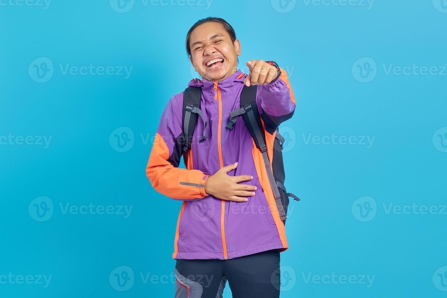 Portrait of handsome Asian young man laughing widely and pointing finger in front isolated on blue background photo