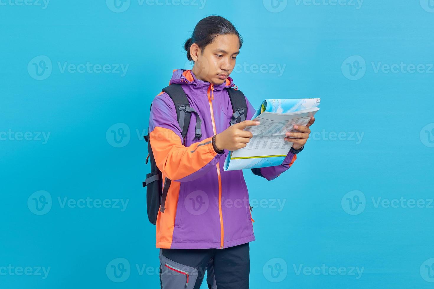 retrato, de, asiático, joven, llevando, chaqueta, y, bolsa de transporte, mirar, serio, lectura, mapa, aislado, en, fondo azul foto