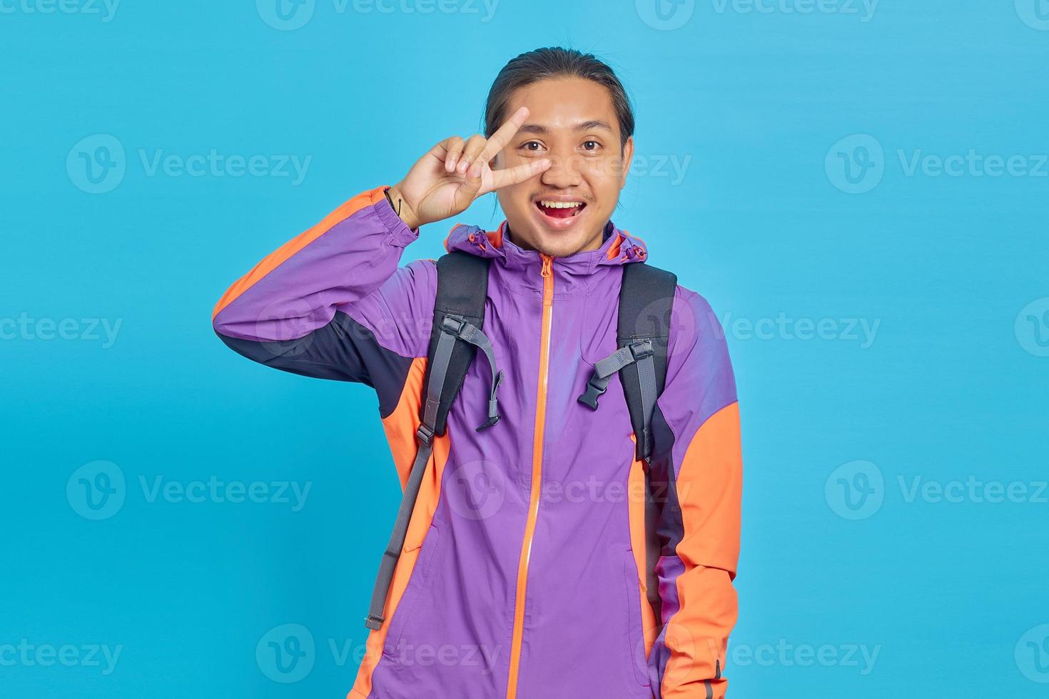 Portrait of smiling young Asian man showing peace gesture and looking at camera isolated on blue background photo