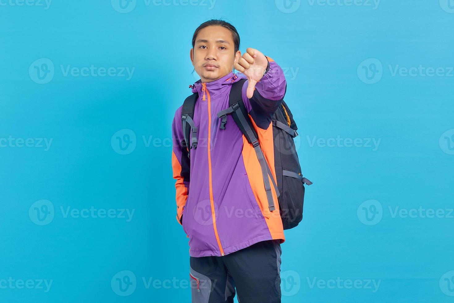 Upset handsome asian man in purple jacket showing thumbs down isolated over blue background photo