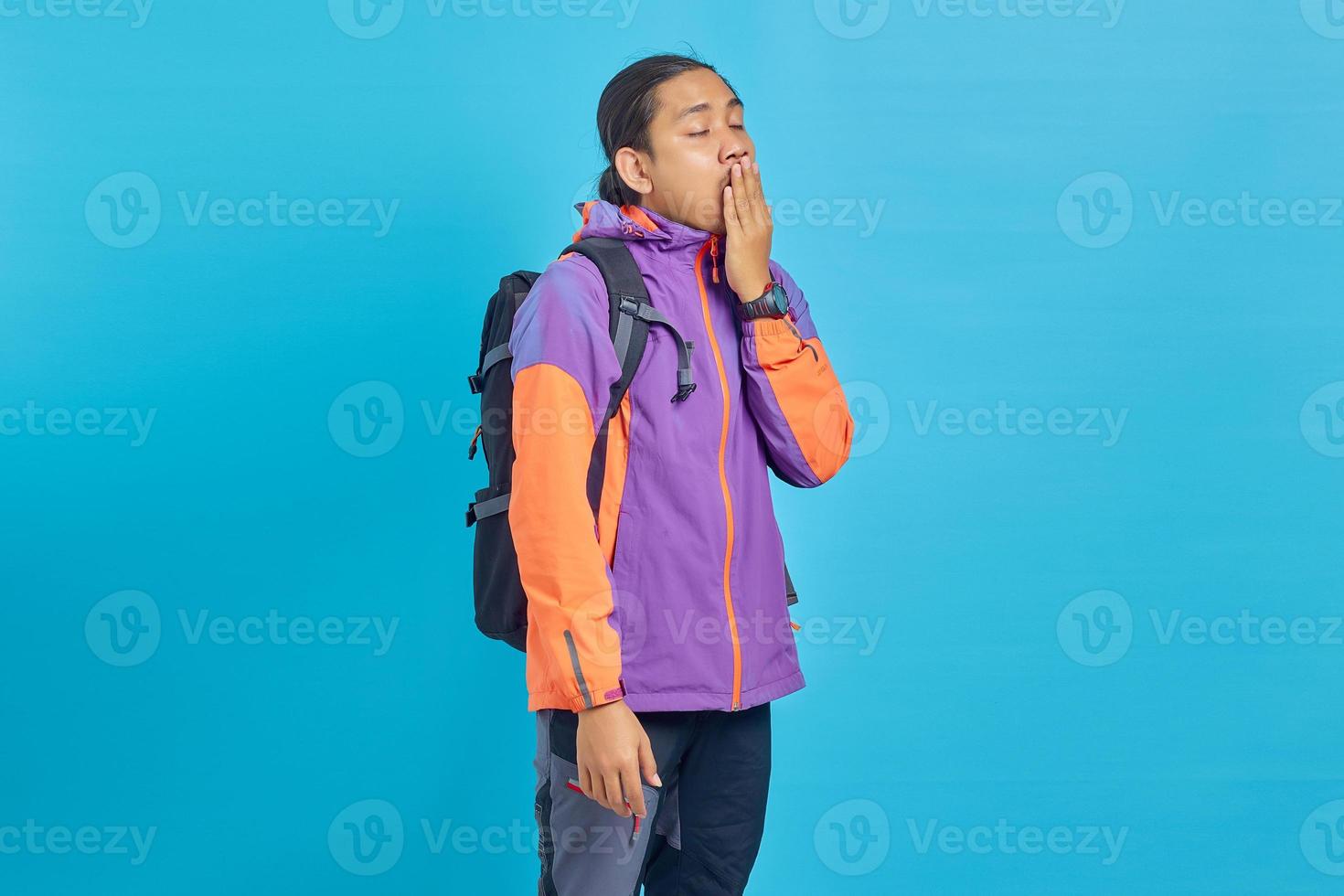 Handsome asian man wearing purple jacket looking sleepy and covering mouth with hands on blue background photo