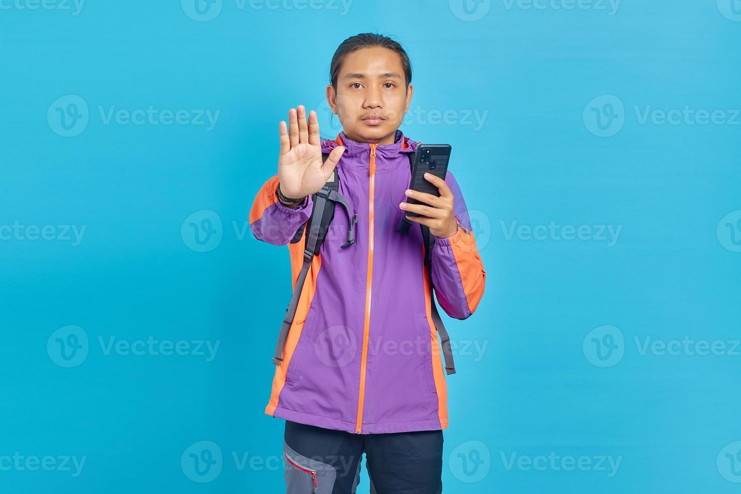 Retrato de hombre asiático joven serio de pie con los brazos extendidos mostrando gesto de parada y sosteniendo el teléfono móvil aislado sobre fondo azul. foto