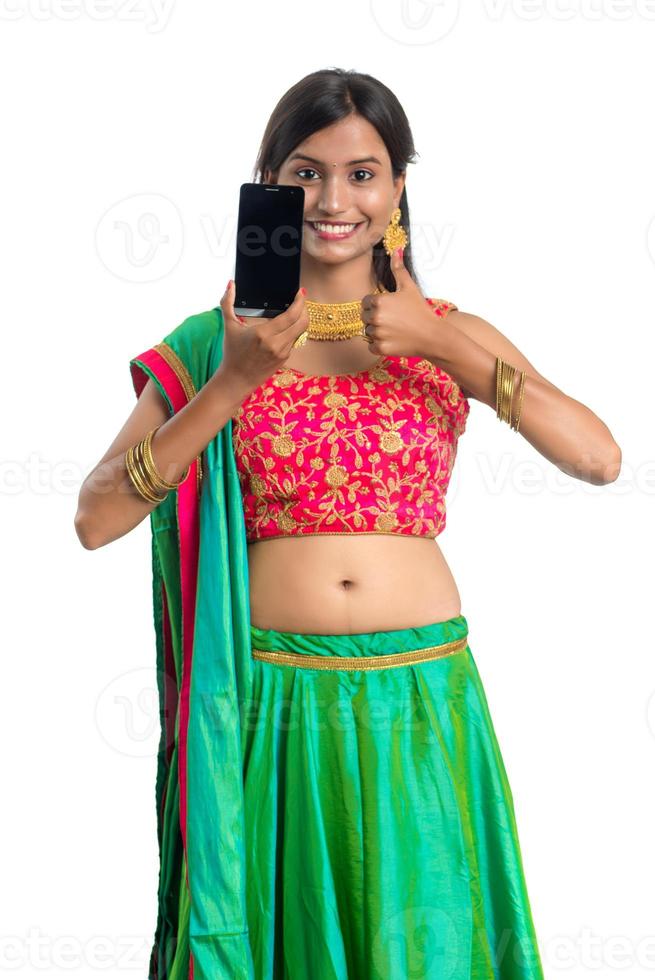 Joven niña tradicional india que usa un teléfono móvil o un teléfono inteligente y muestra un teléfono inteligente con pantalla en blanco sobre fondo blanco. foto