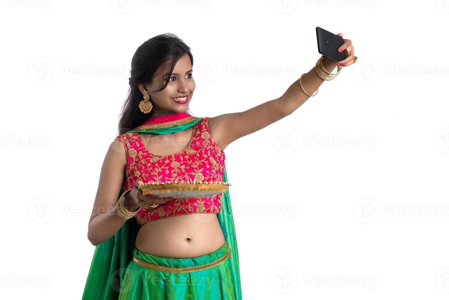 Hermosa joven feliz tomando un selfie con lámpara de arcilla o diya durante el festival de diwali ligero usando un teléfono inteligente sobre un fondo blanco. foto