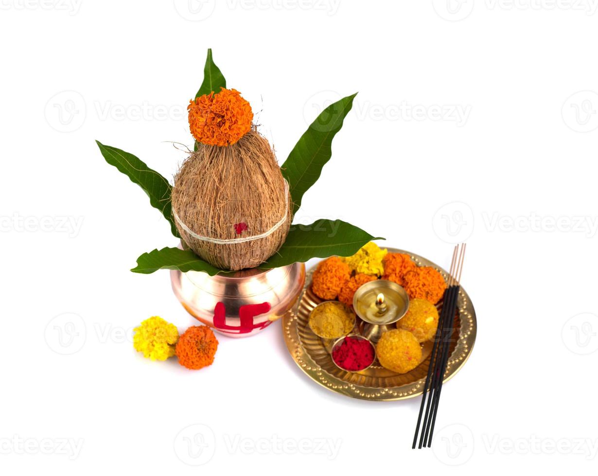 kalash de cobre con hoja de coco y mango y pooja thali con diya, kumkum y dulces con decoración floral sobre fondo blanco. esencial en puja hindú. foto