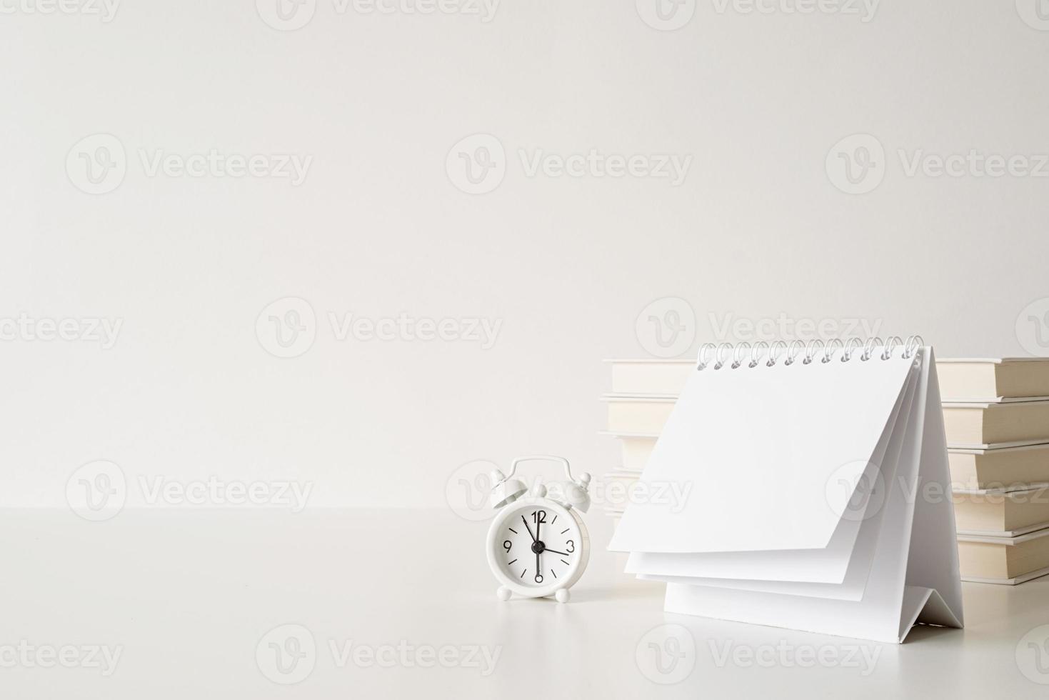 Simulacros de calendario en espiral en blanco con hojas volteadas sobre una mesa blanca con pila de libros, vasos y reloj foto