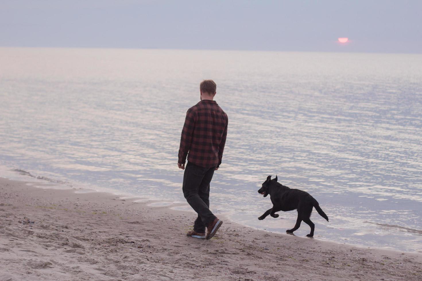 Joven caminando por la playa por la mañana con perro negro foto