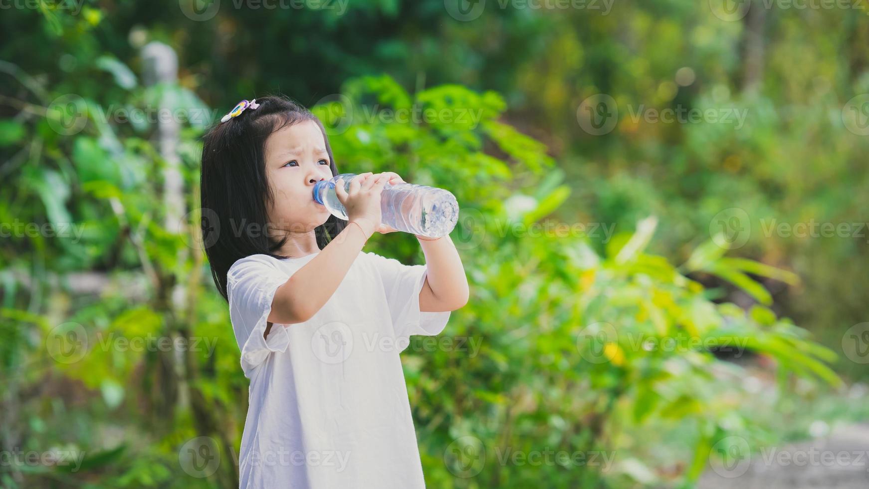 4 years Asian kid drinking water from plastic bottle. Child girl was thirsty. Green nature background. Empty space to enter text. photo