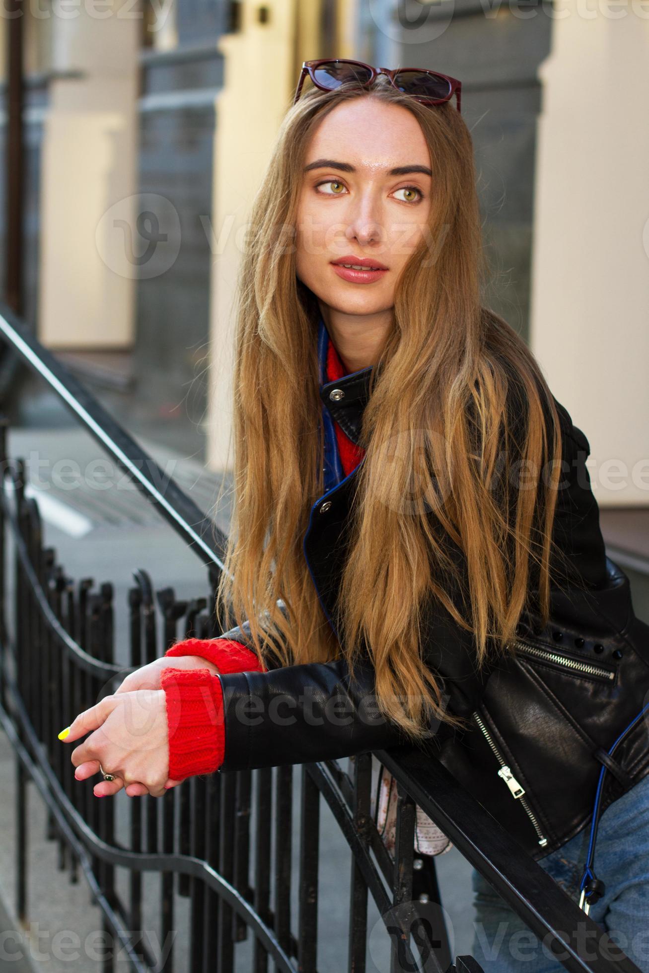 Cool bastante joven zoomer mujer con chaqueta blanca de moda de cuero y  jeans usa unas elegantes gafas de sol se sienta en los escalones y descansa  cerca de un edificio de