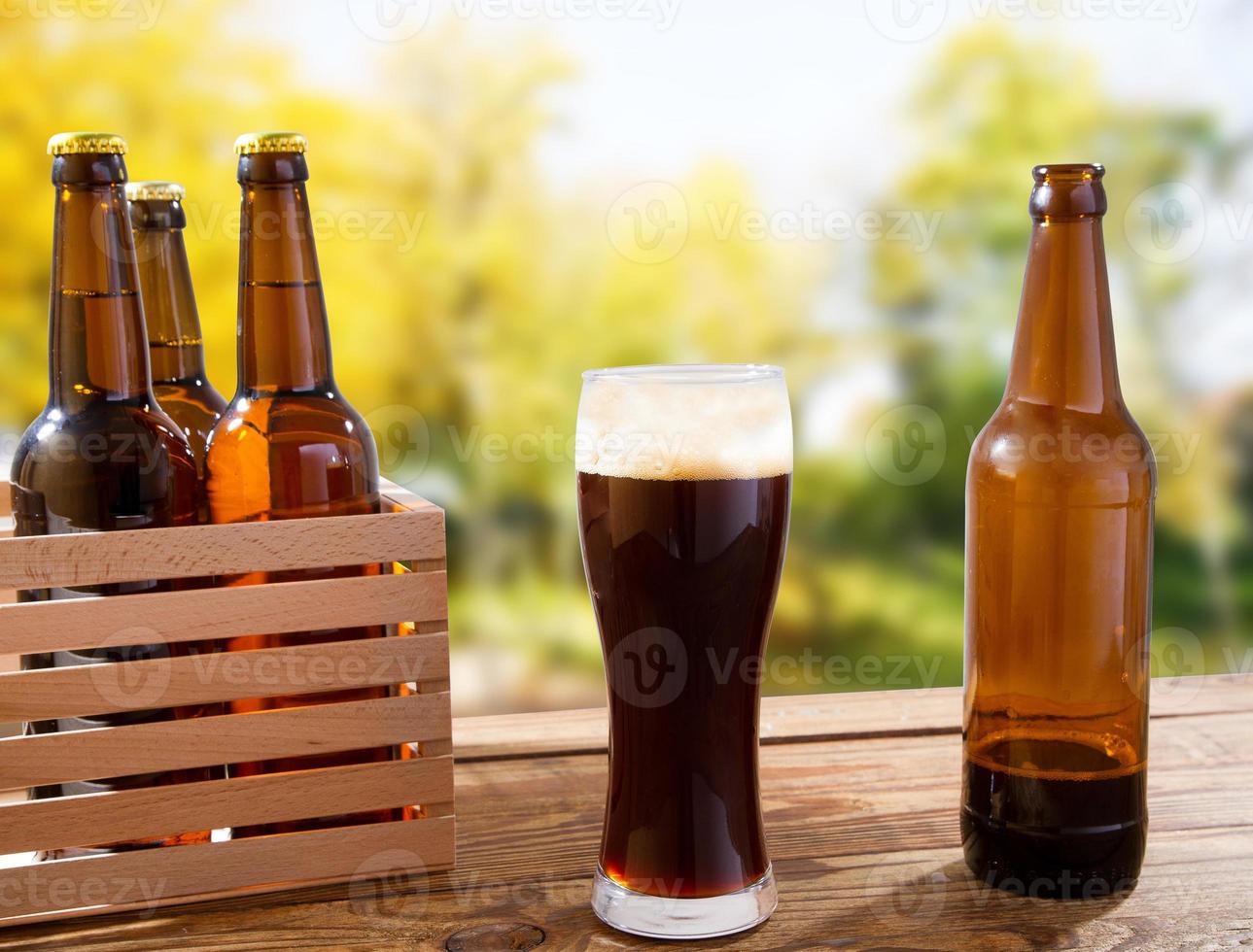 cup of dark beer and bottles on wooden box on table on blurred park background photo