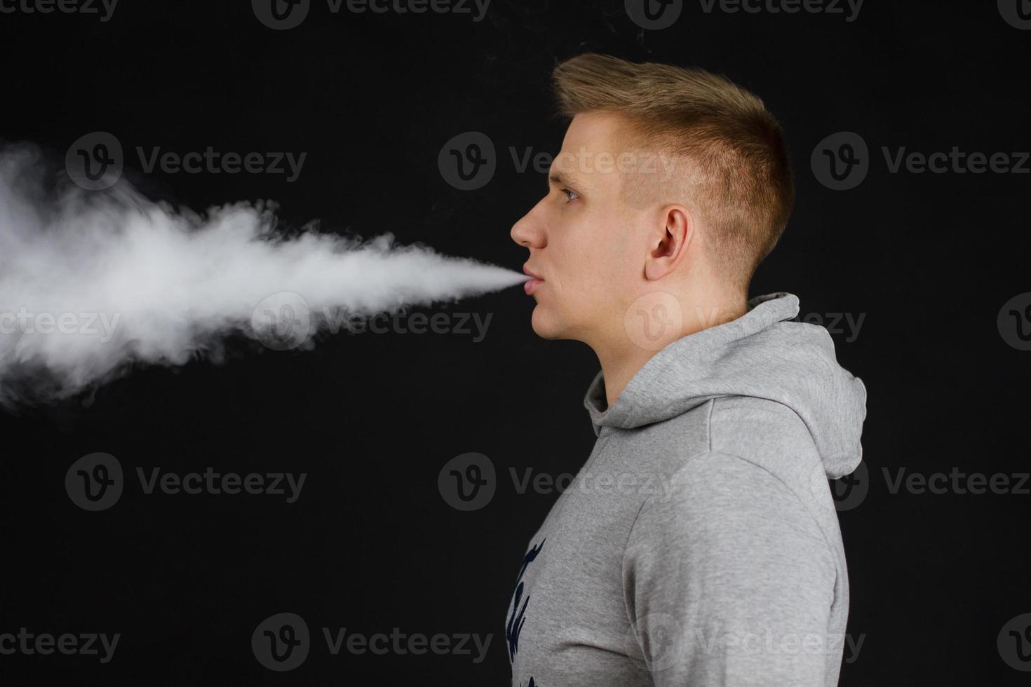Men in hoodie vaping and releases a cloud of vapor. Selective focus photo