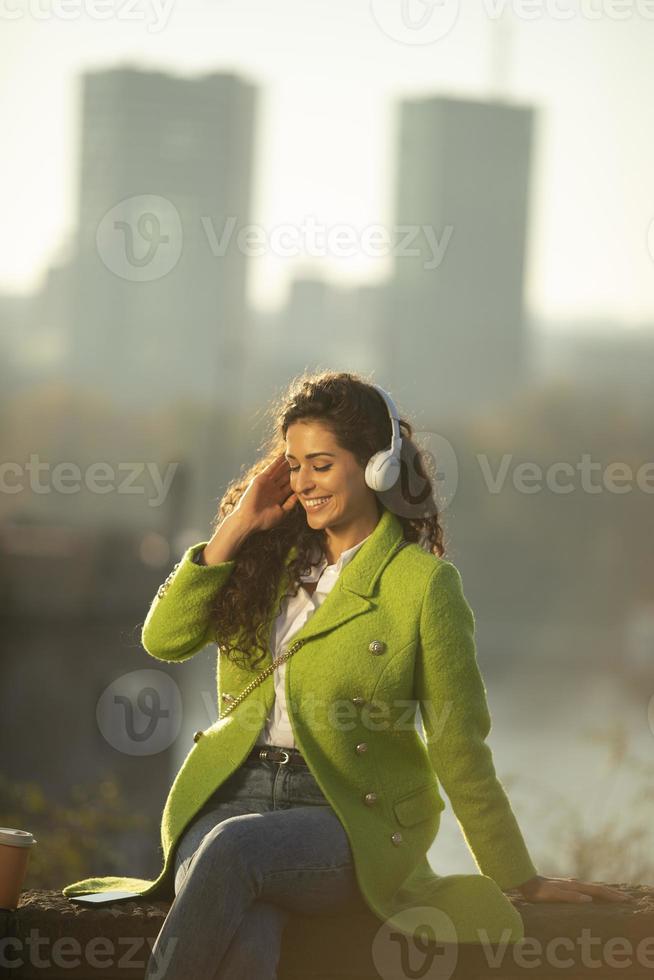 Pretty young woman listening music with smartphone and takeaway coffee by the river on a sunny autumn day photo