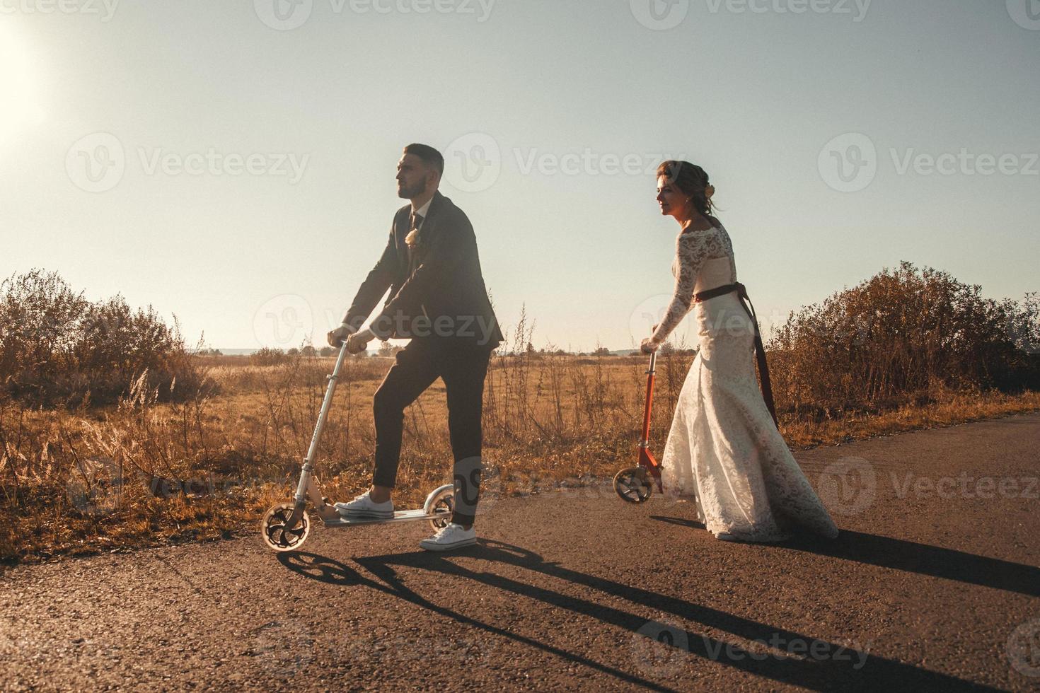 Sonriente pareja de novios montando en scooters a lo largo de la carretera fuera de la ciudad al atardecer. lugar para texto o publicidad foto