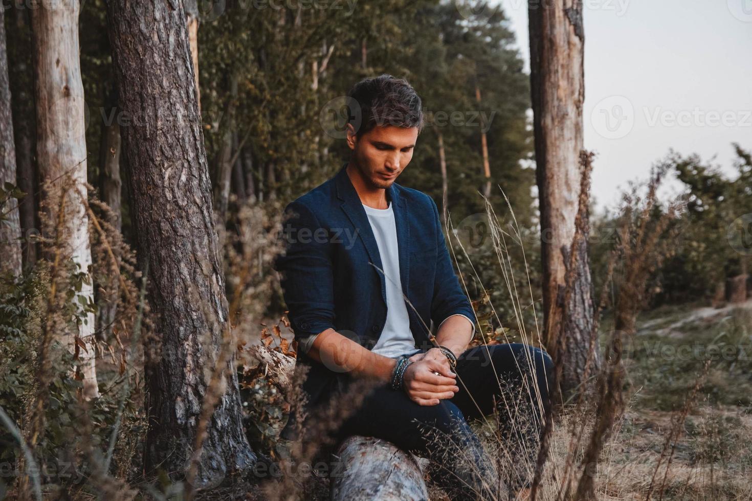 Handsome man posing outdoors in forest sits on a log, wearing checked jacket. Place for text or advertising photo