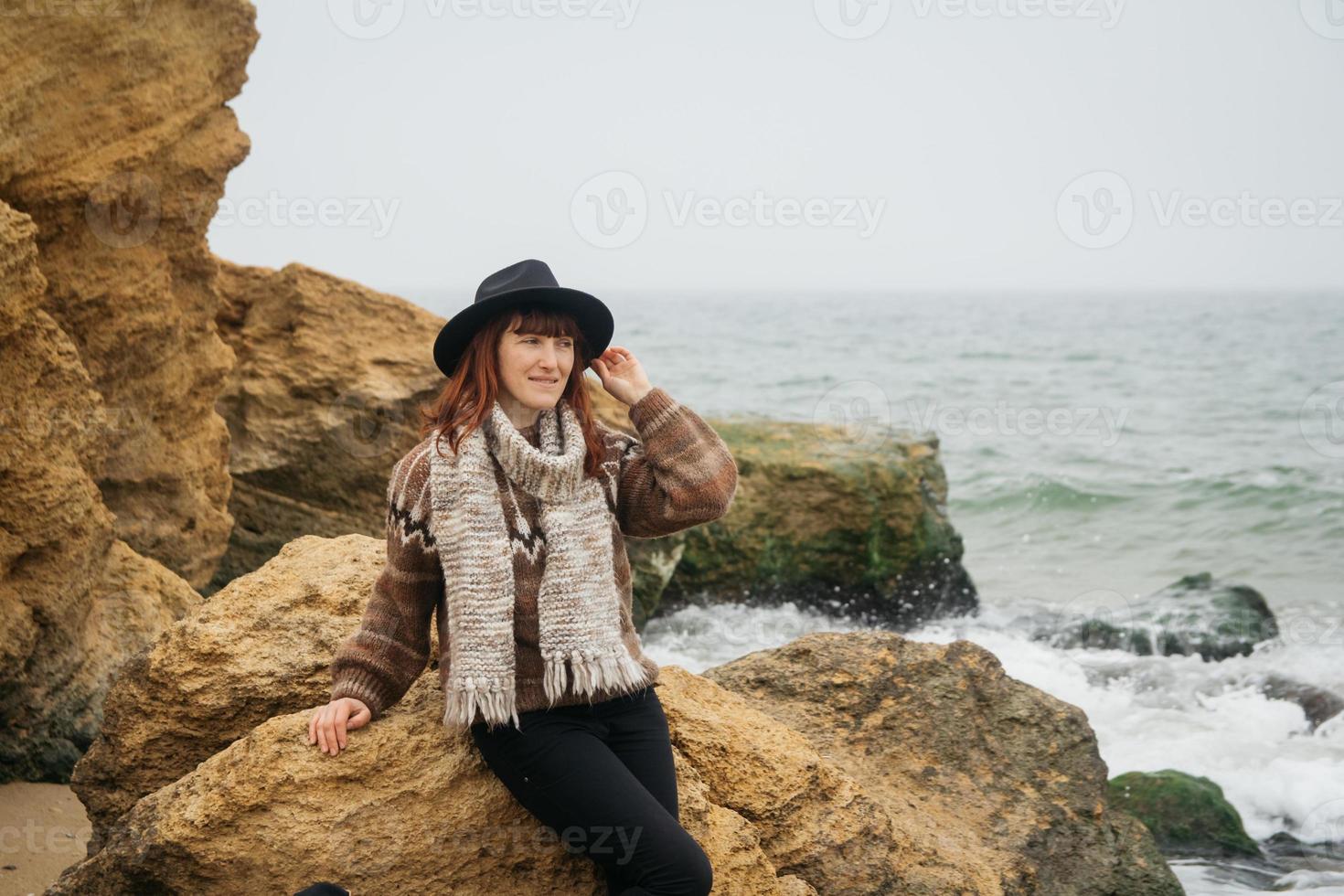 Mujer con sombrero y bufanda de pie sobre una roca frente a un hermoso mar en el horizonte foto