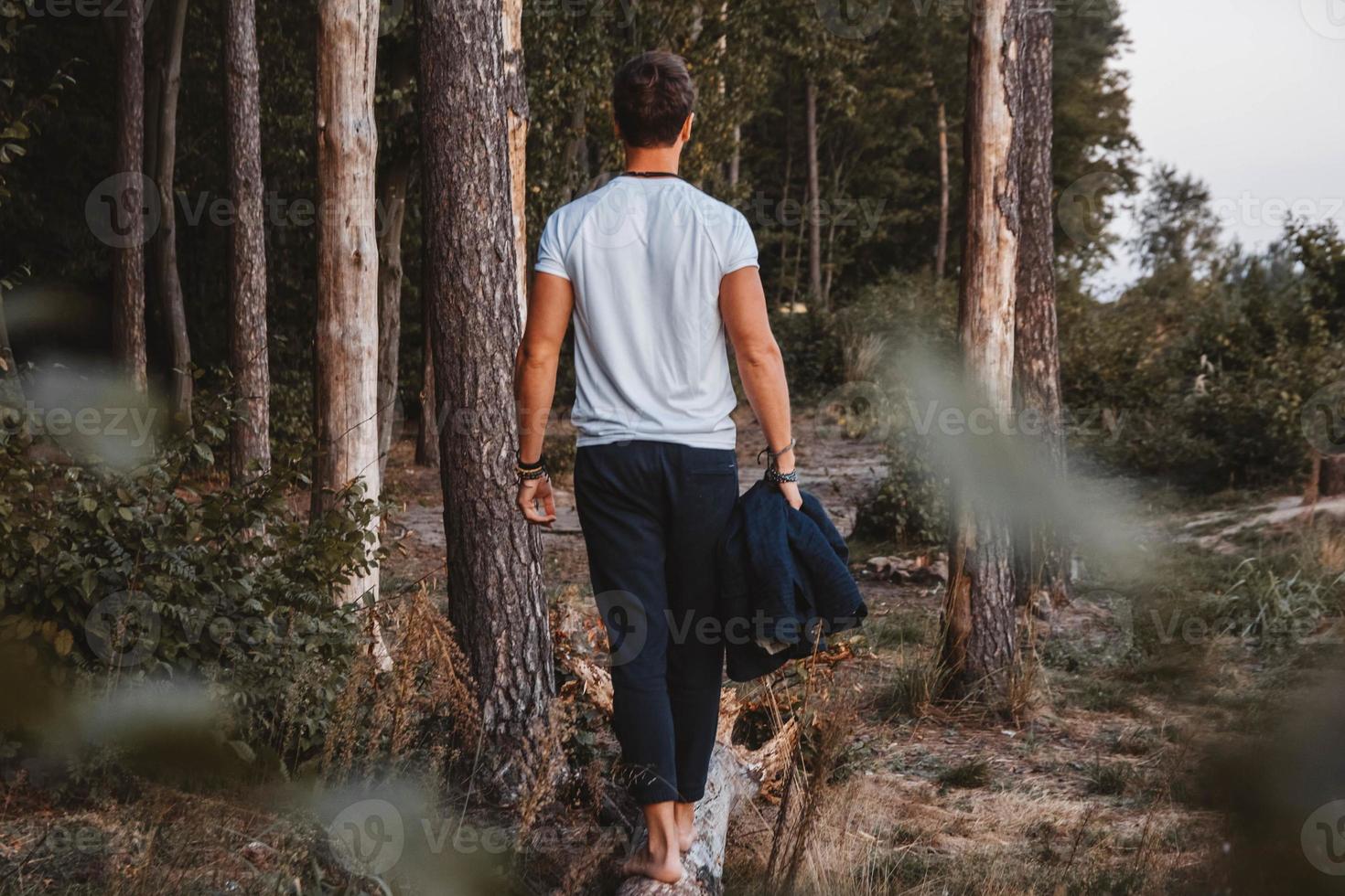 Handsome man posing outdoors in forest stands barefoot on a log, wearing checked jacket. Place for text or advertising photo