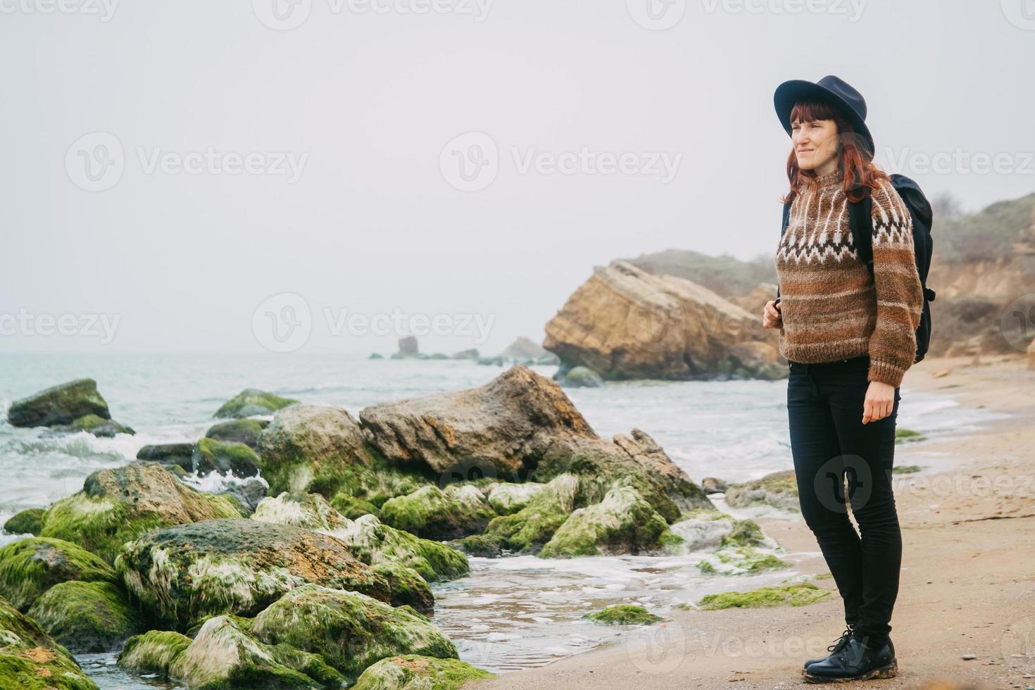 Woman in a hat and scarf with a backpack on coast against background of rocks against beautiful sea photo