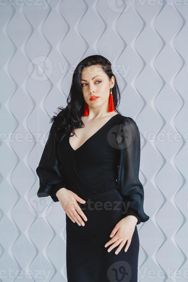 Fashion portrait of a stylish brunette woman wearing a black dress. Woman with long hair wearing red earrings. Posing in studio. Place for text or advertising photo