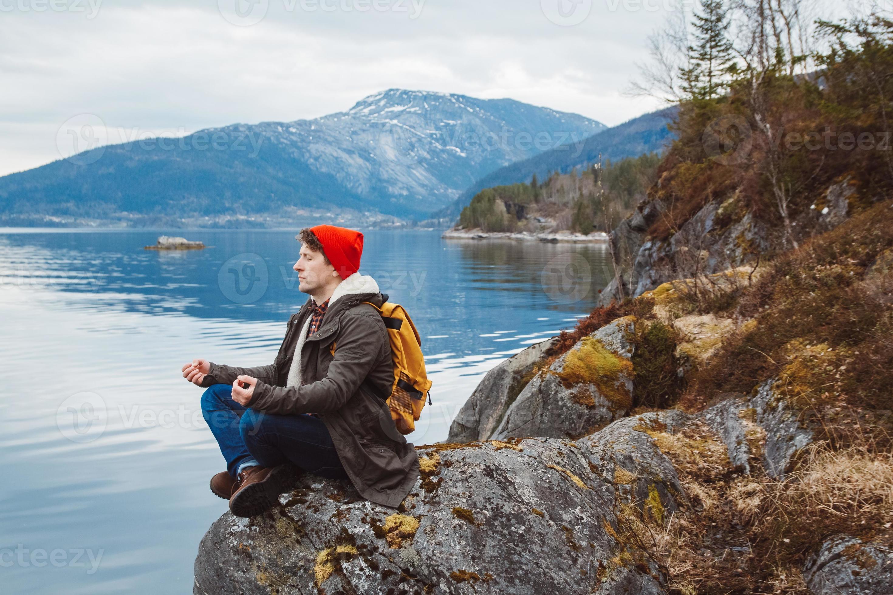 Traveler man in a meditative position sitting on a rocky shore on the background of a mountain and a lake. Space for your text message or promotional content photo