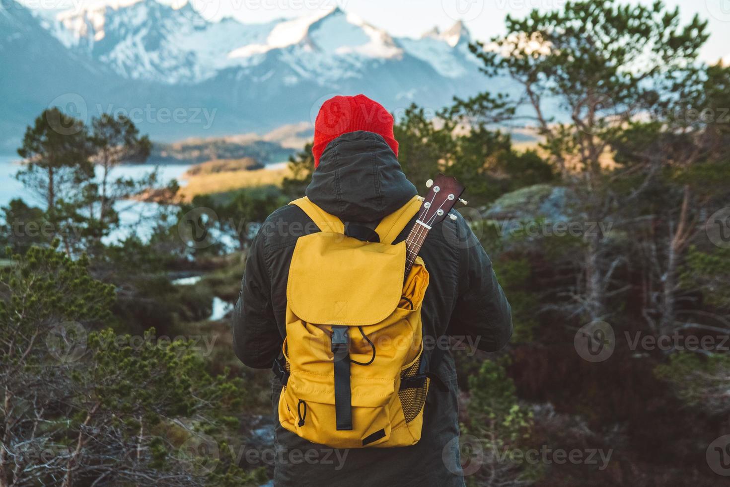 Traveler man is standing in the middle of a forest with a guitar on background of mountains and lake. Wearing a yellow backpack in a red hat. Place for text or advertising. Shoot from the back photo