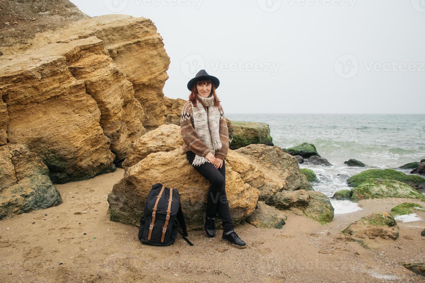 Mujer con sombrero y bufanda con una mochila en la costa con el telón de fondo de las rocas contra el hermoso mar foto