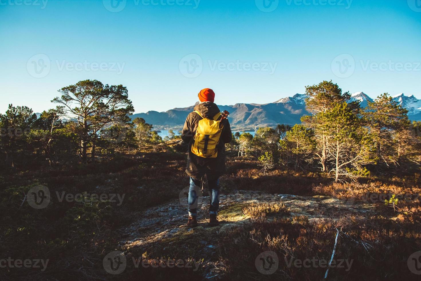 Man is standing in the middle of a forest with a yellow backpack and guitar on background of mountains and lake photo